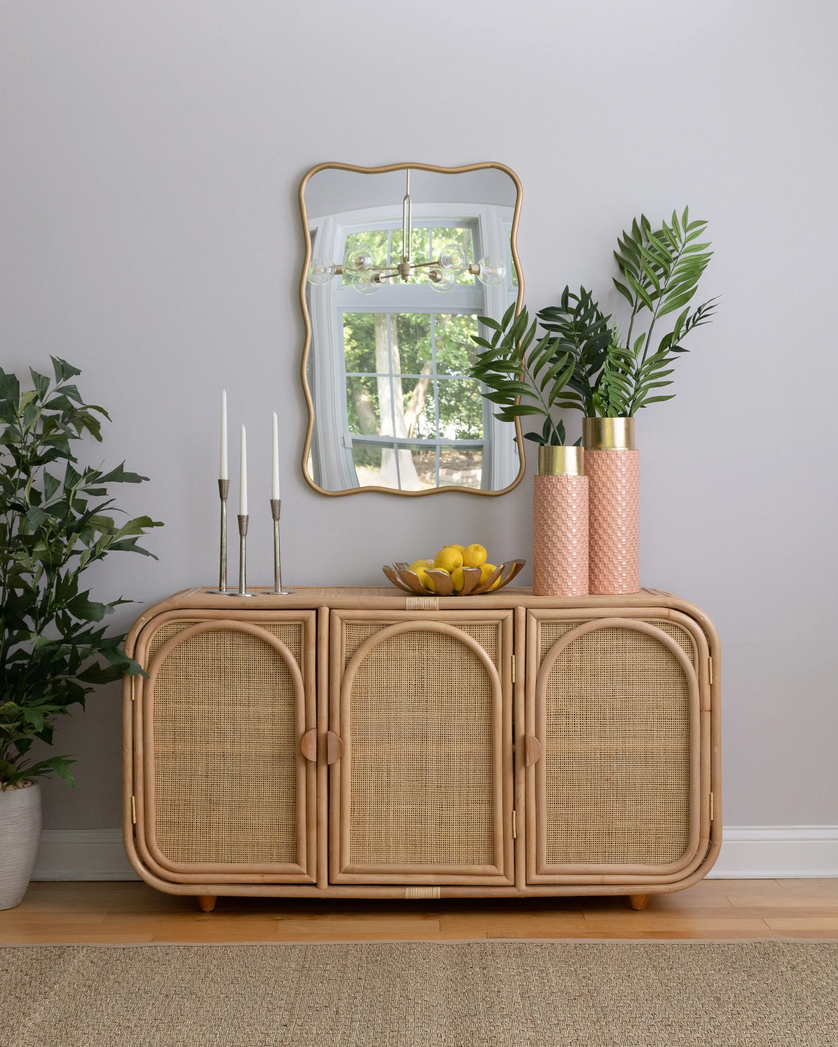 A dining room styled with natural, earthy tones and textures. On the left, a wooden rattan console table is adorned with tall, slim candles, leafy plants in decorative vases, and a large gold-framed mirror above. In the center, a vibrant green indoor plant stands in a woven basket, adding height and life to the corner. On the right, a simple wooden dining table is paired with woven rattan chairs, set against large windows that flood the room with natural light. The space feels warm and inviting, with a focus on natural materials and serene, neutral decor.