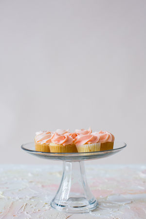 A glass cake stand with a row of soft pink cupcakes topped with swirled buttercream frosting, creating a minimalistic and elegant dessert display.