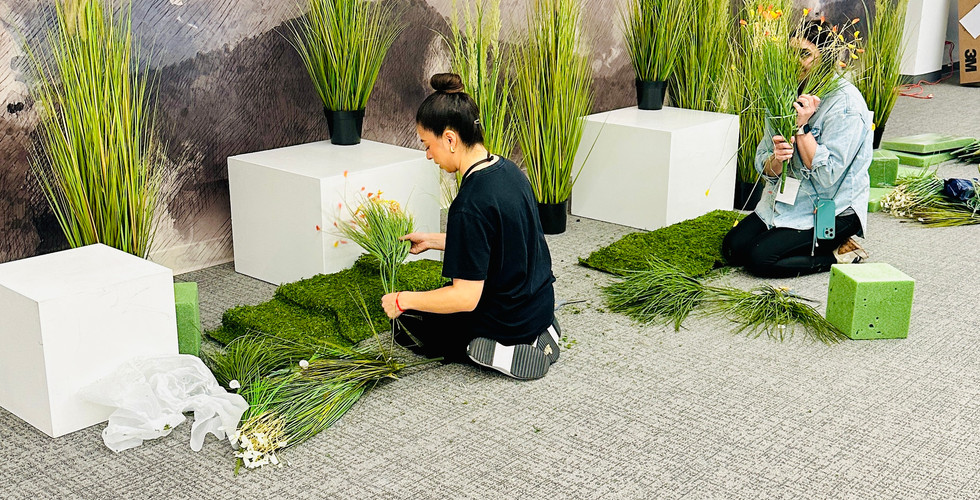 Two stylists on the floor arranging faux greenery and florals at the base of a mural wall for the National Bridal Market Chicago display.