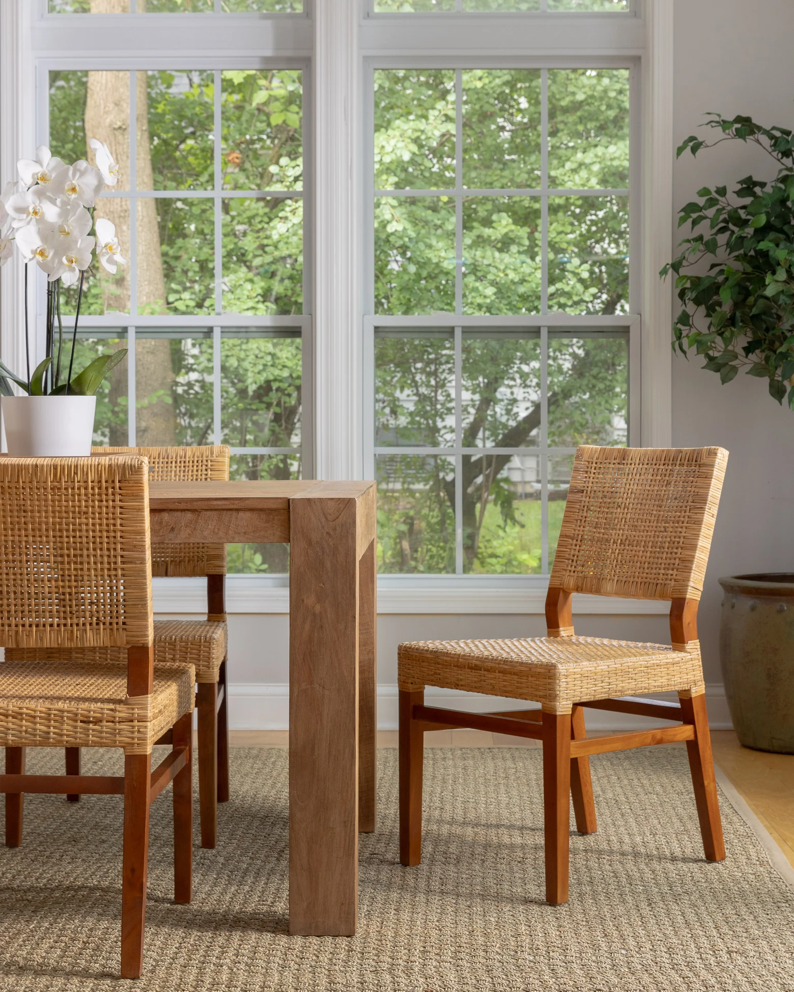 A dining room styled with natural, earthy tones and textures. On the left, a wooden rattan console table is adorned with tall, slim candles, leafy plants in decorative vases, and a large gold-framed mirror above. In the center, a vibrant green indoor plant stands in a woven basket, adding height and life to the corner. On the right, a simple wooden dining table is paired with woven rattan chairs, set against large windows that flood the room with natural light. The space feels warm and inviting, with a focus on natural materials and serene, neutral decor.
