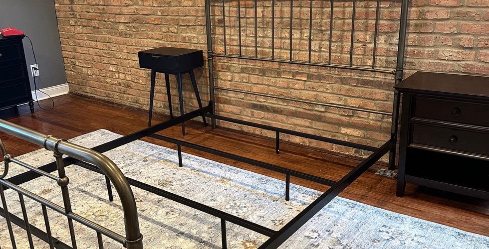 A black metal bed frame partially assembled in a bedroom with a brick accent wall and a light gray area rug.