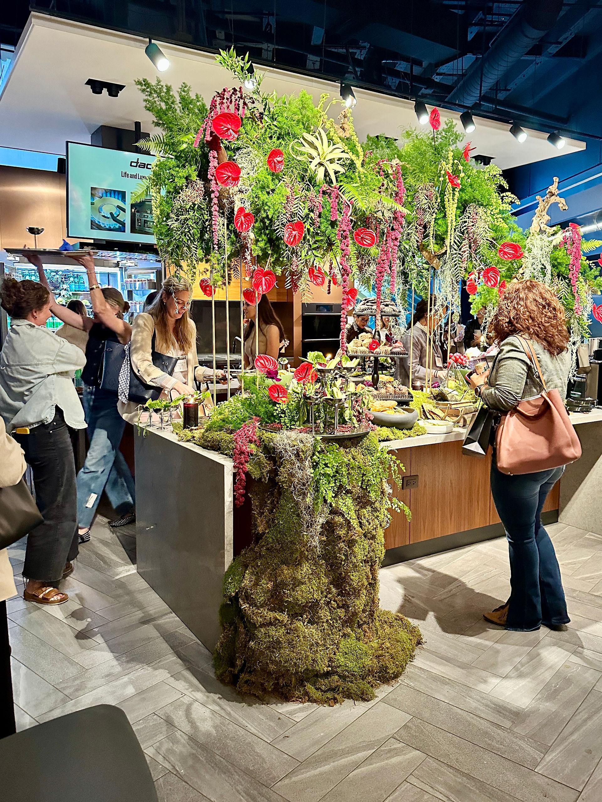 A wide shot of an elaborate tablescape installation with tall, lush greenery and florals hanging above a food display, as attendees admire the striking setup at a design event.