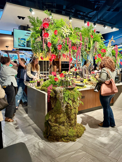 A wide shot of an elaborate tablescape installation with tall, lush greenery and florals hanging above a food display, as attendees admire the striking setup at a design event.