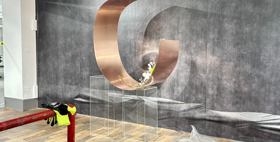 Early stage of floral display setup showing a copper crescent structure on a dark wall, with tools and materials on the ground.