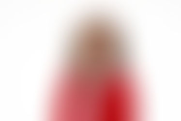 Portrait of a smiling woman with curly hair, red glasses, and a red jacket over a white collared shirt, against a plain white background.