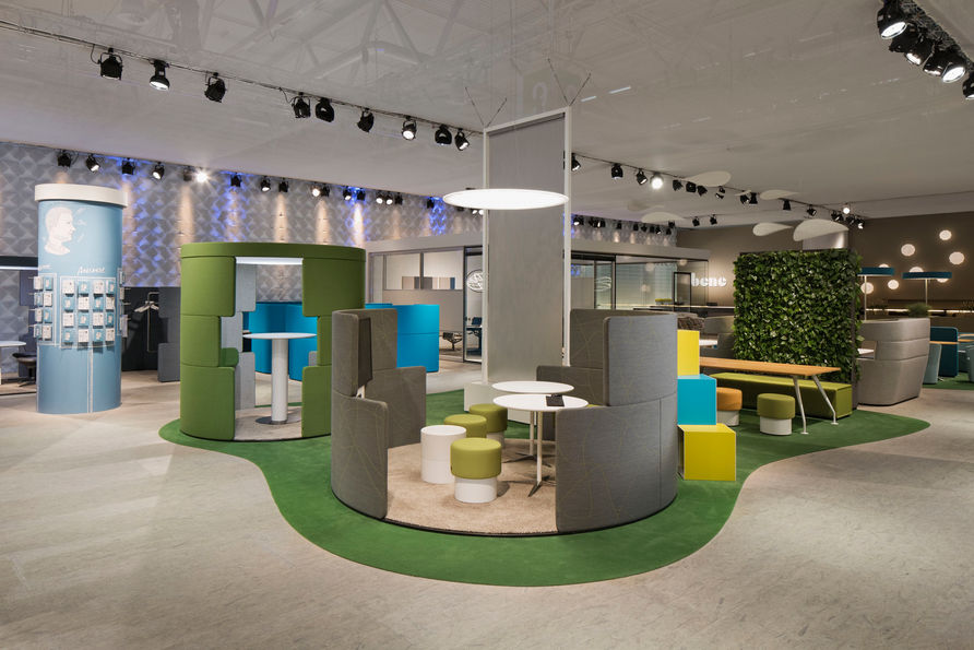 一個寬敞的展廳或辦公空間，地面鋪著綠色地毯，上面擺放著各種色彩鮮豔的辦公家具，包括獨立的工作艙和組合式沙發，呈現出一個充滿活力和創意的環境。