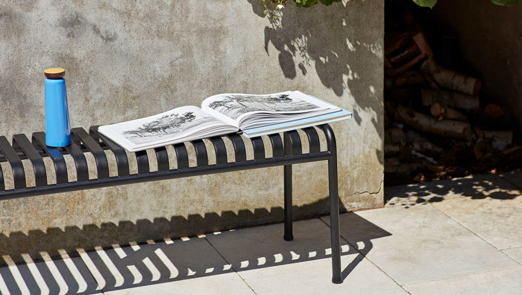 PALISSADE格柵長凳上放著一本打開的書和一個藍色水瓶，椅子投射條紋陰影，背景為混凝土牆和一些樹枝影。