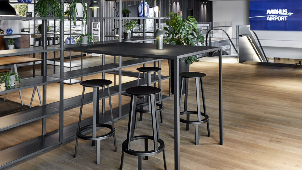 現代機場候機區,黑色REVOLVER高桌椅,桌上擺綠色植物,背景為木地板和牆上“AARHUS AIRPORT”標誌。