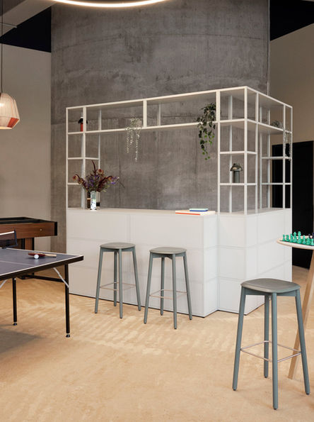 開放式室內環境,灰色背景牆前有白色吧台,擺放綠色植物和書籍、SOFT EDGE 木椅凳。前景有乒乓桌,氣氛簡約寧靜。