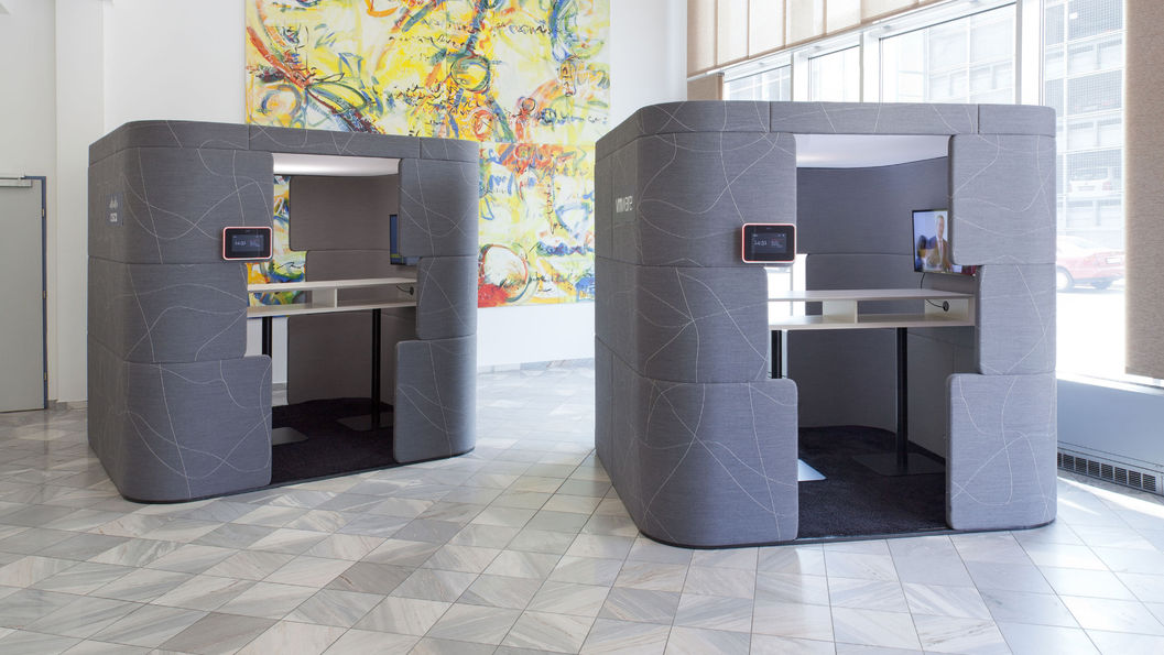 這張照片展示了兩款灰色的隔音工作艙，內部都配有桌子和螢幕，可以作為獨立的辦公或會議空間。