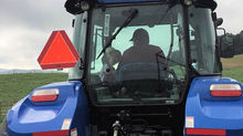 Rear view of blue tractor with driver silhouette, orange slow-moving triangle, and Augusta Equipment label on a cloudy field.