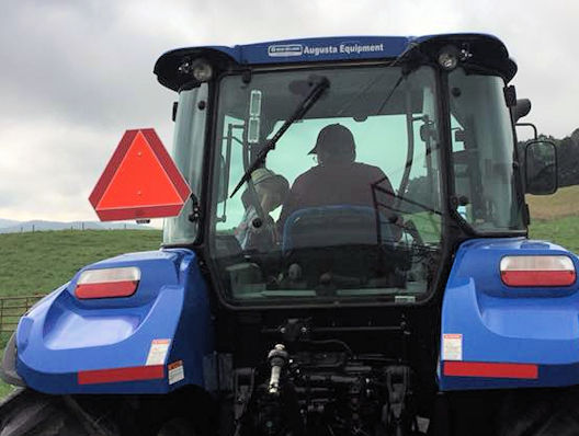 Rear view of blue tractor with driver silhouette, orange slow-moving triangle, and Augusta Equipment label on a cloudy field.
