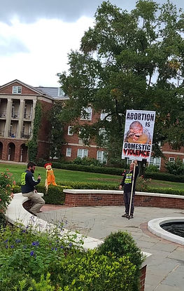 One man sitting in the courtyard, another holding a sign that says ‘Abortion is Domestic Violence’ with a small baby on it.