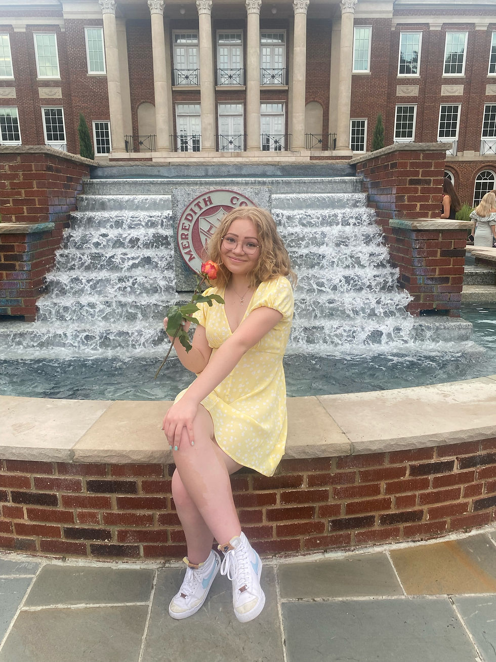 A girl in a yellow dress holding a pink rose and sitting on the Meredith fountain with the Lux symbol behind it