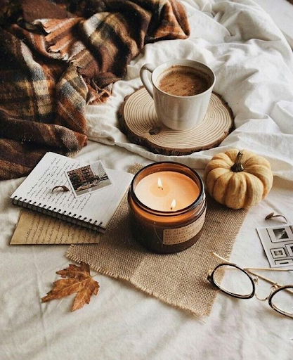 A cup of coffee, lit candle, small pumpkin, and journal on a bed with fall leaves scattered around and a cozy blanket to the side