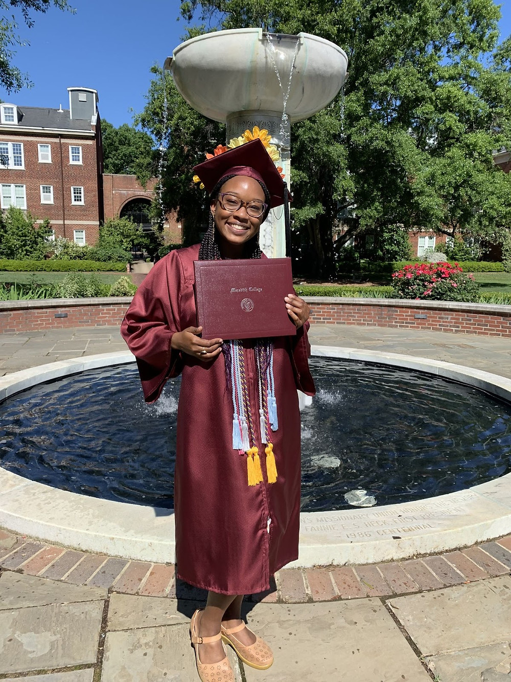 Shila standing by a fountain on Meredith's campus in her cap and gown, holding her diploma