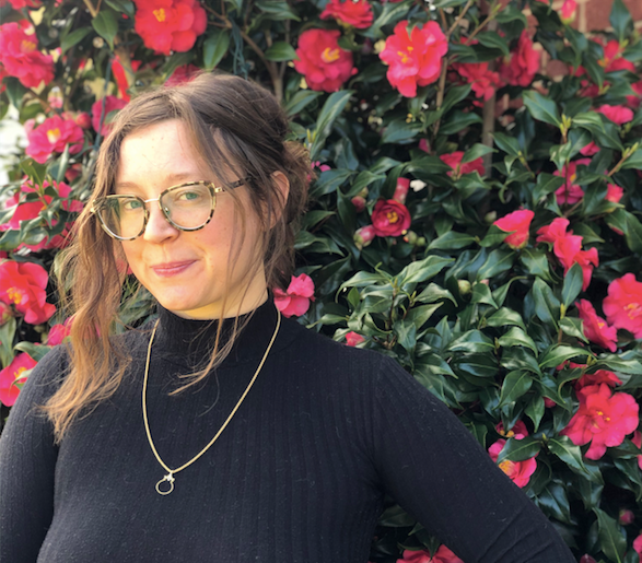 Chanelle, a brunette woman wearing glasses, standing in front of a pink flowering bush wearing a black turtleneck