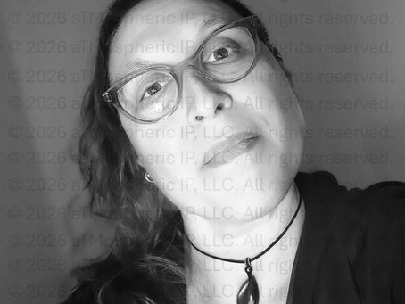 Selfie of a woman with wavy hair and glasses, looking down at the camera. She wears a dark choker necklace with a teardrop-shaped stone pendant, and a dark cardigan. Plain gray background.