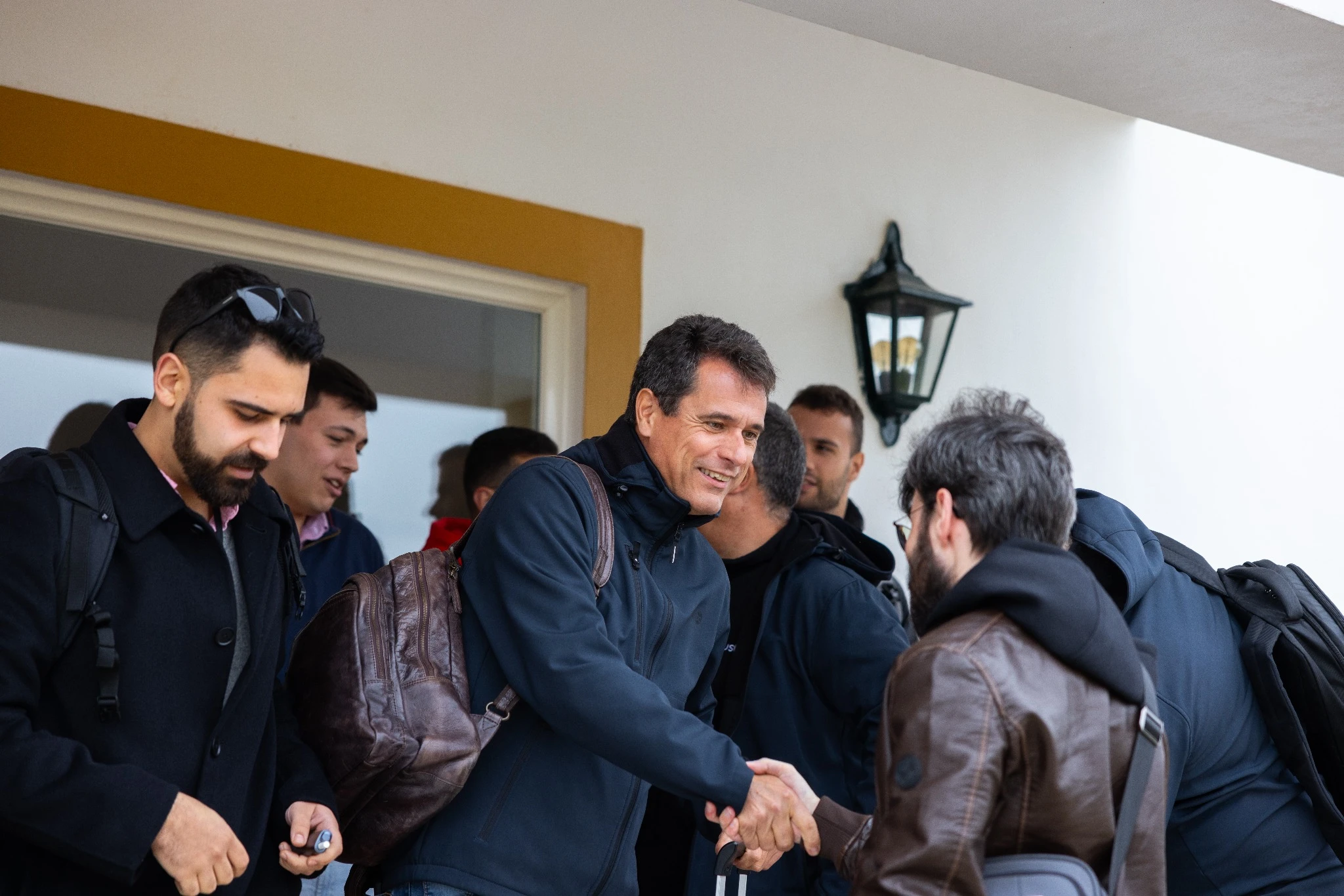 Two colleagues greeting each other with a handshake at the entrance of a venue, while other team members gather in the background.