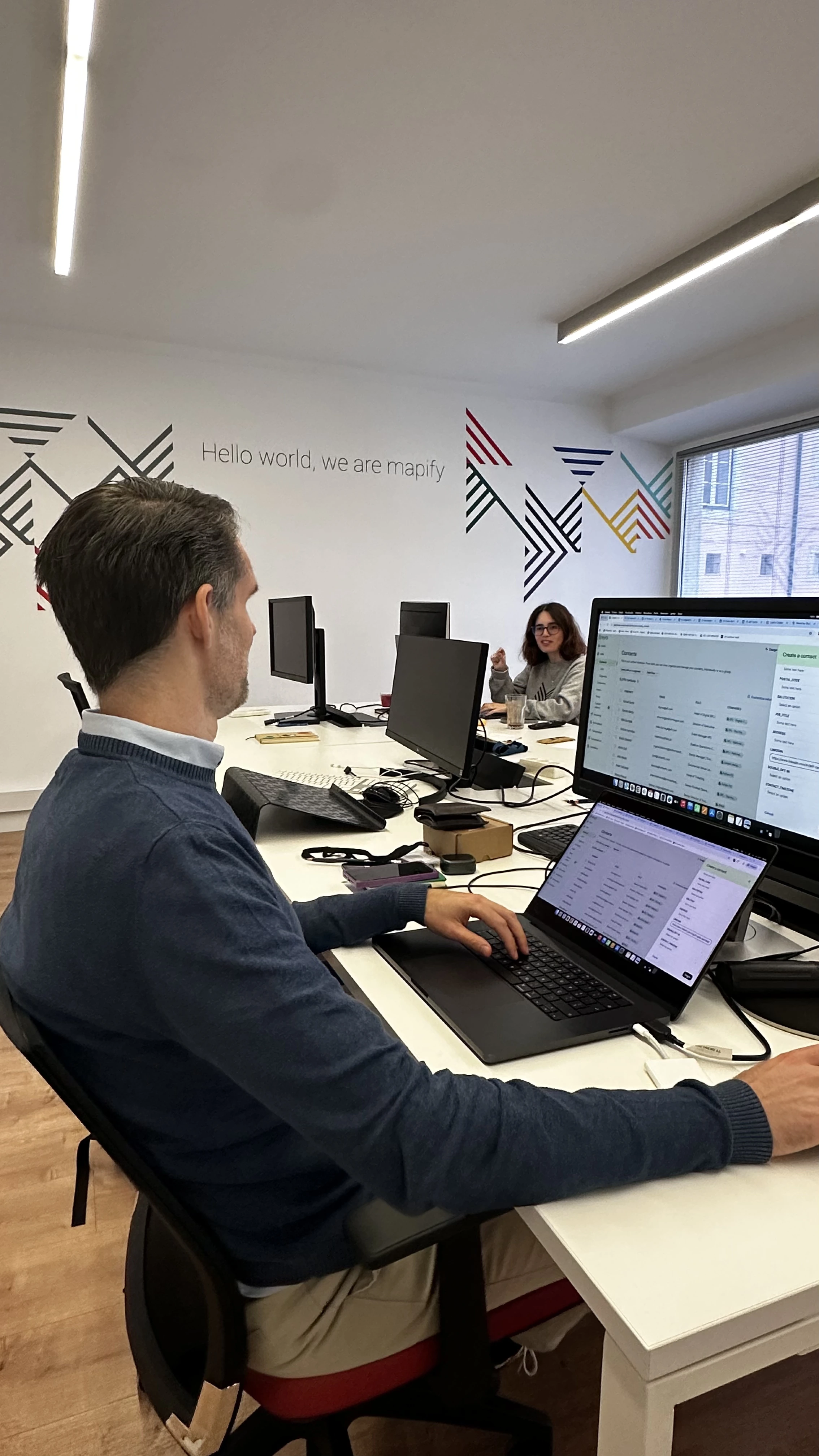A man sitting at his workstation in a modern office, working on a laptop connected to multiple screens. On the wall behind him, the text reads “Hello world, we are mapify.” Another person is seated across the table, engaging in conversation.