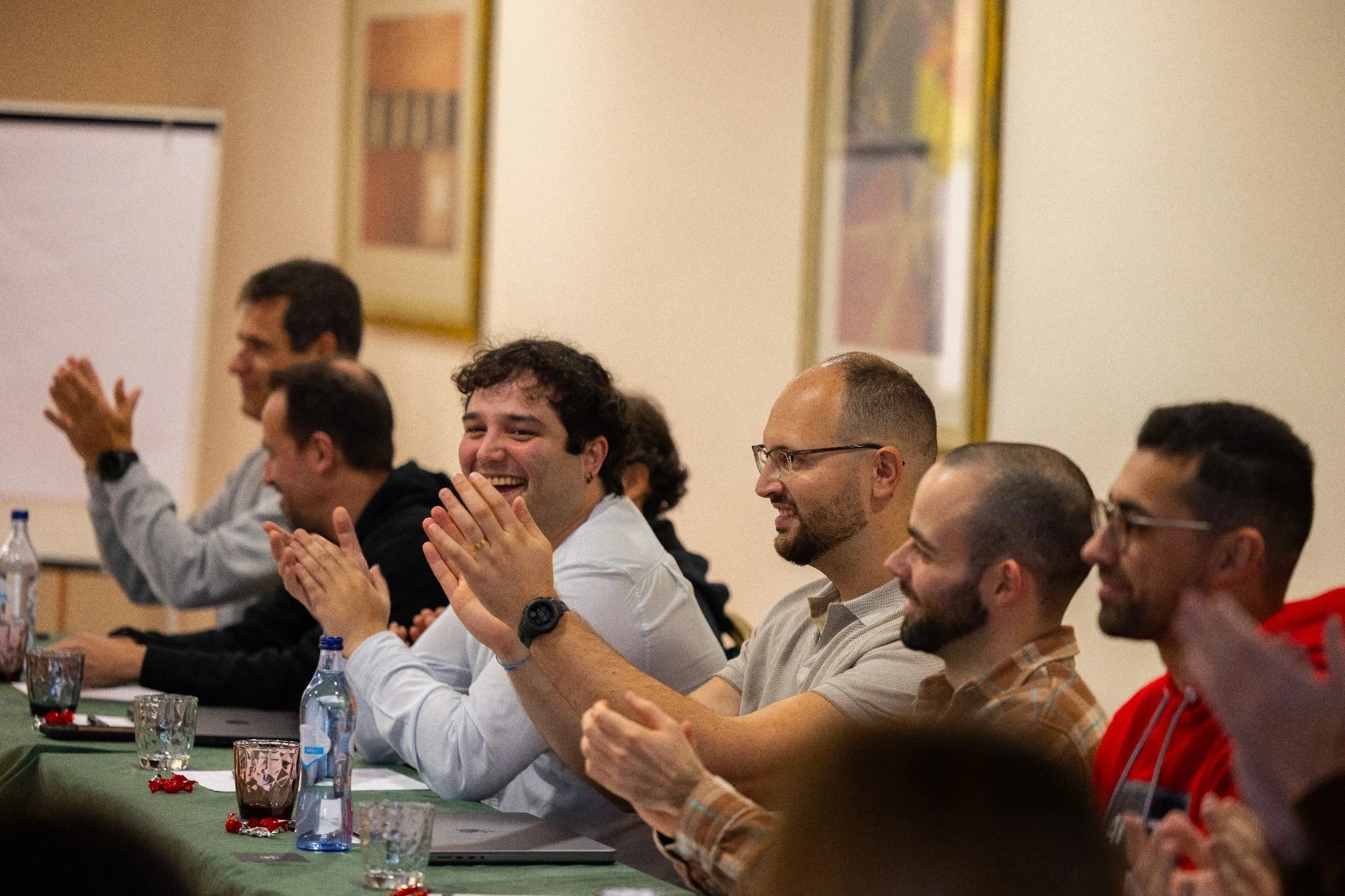Team members seated at a table applauding and smiling during a meeting or presentation, with notebooks and water glasses in front of them.
