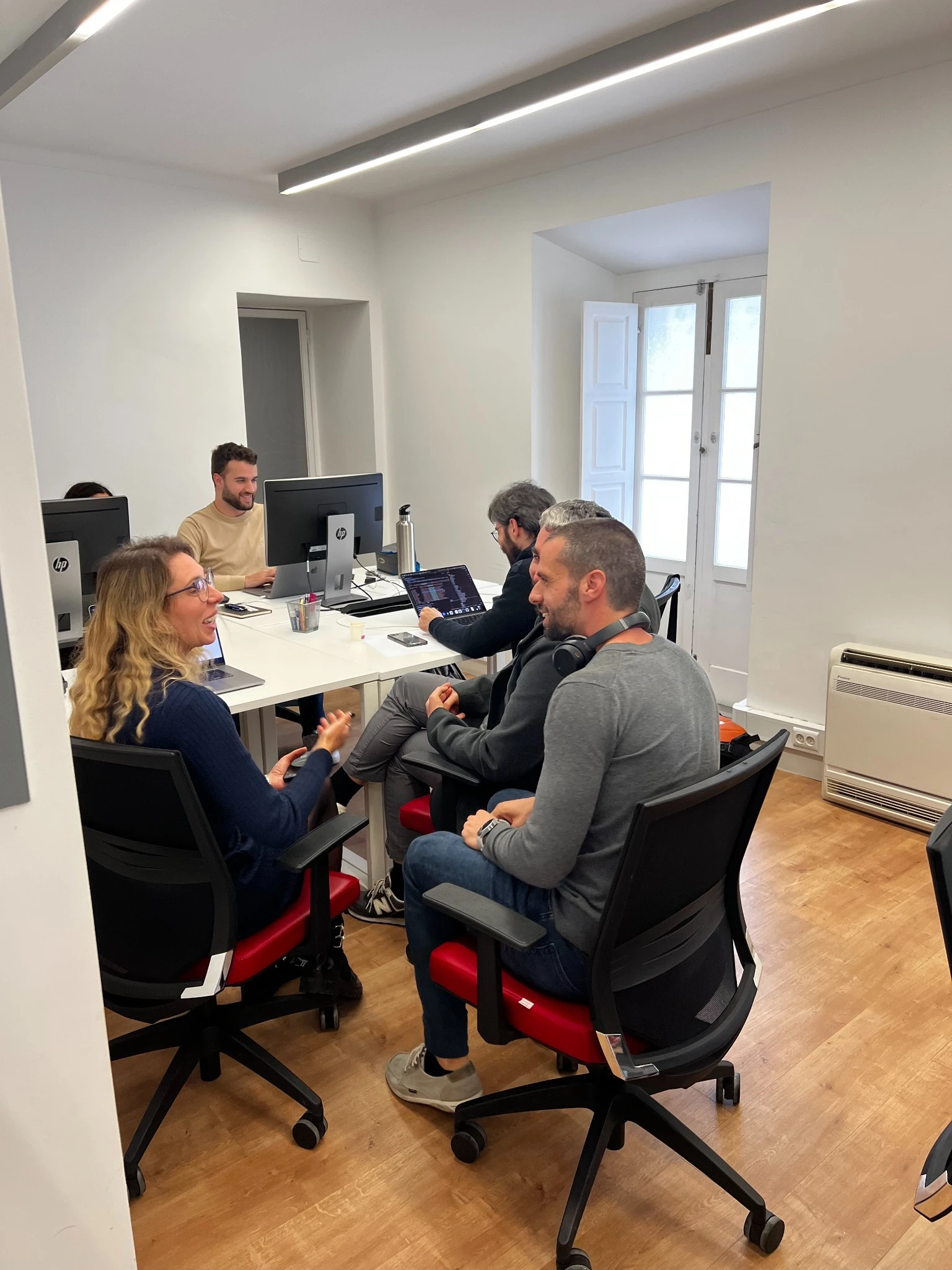 Team members collaborating around a desk in a bright office, working on desktop computers and laptops while discussing ideas.