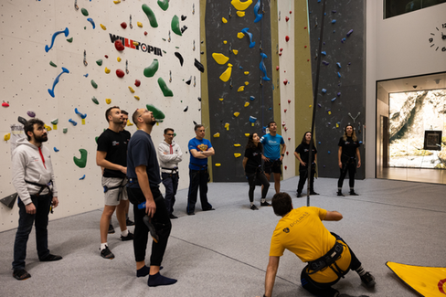 Group gathered on the padded floor looking up at a climber, with ropes and harnesses visible in the climbing area.