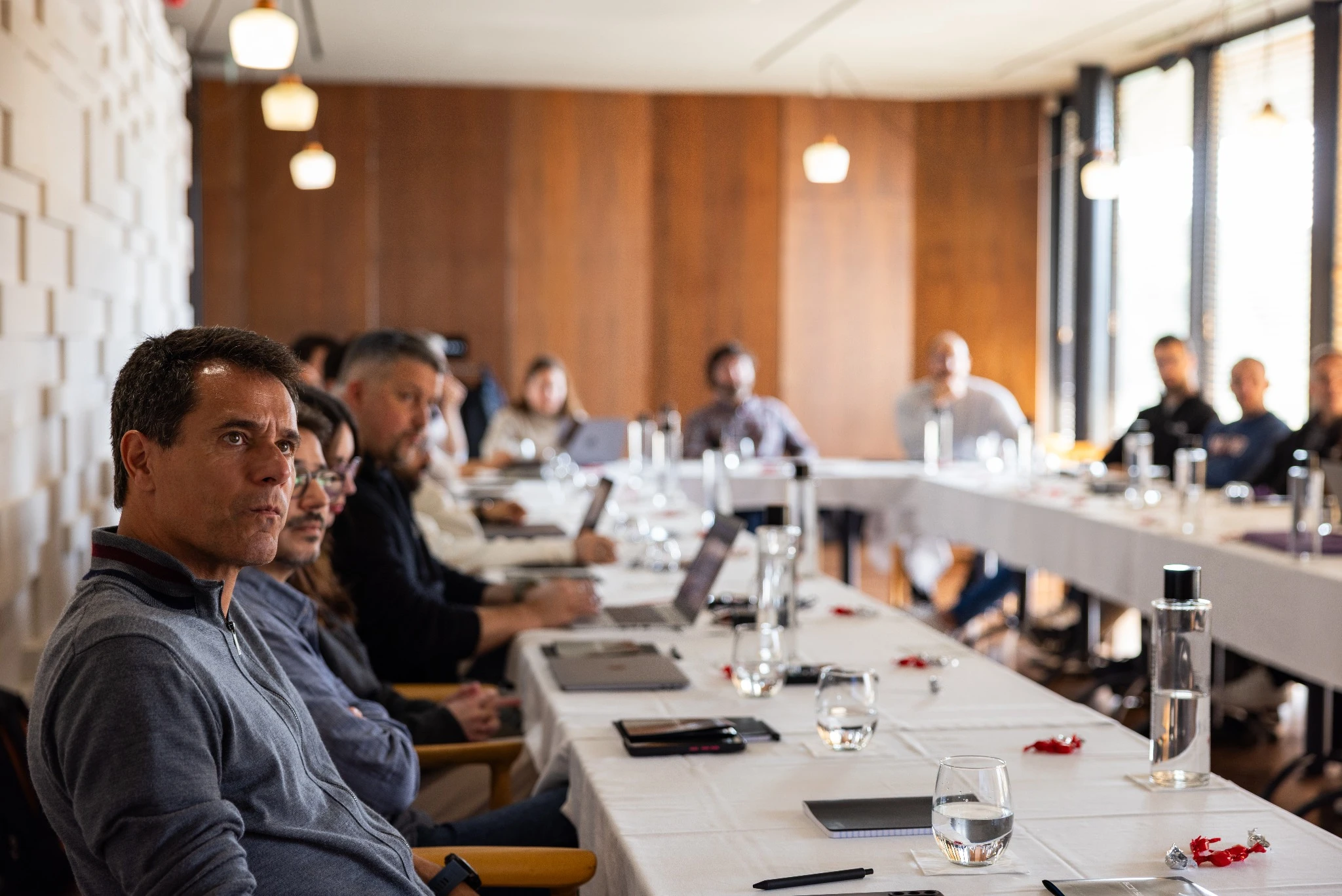 Team members seated around a U-shaped table during a meeting, listening attentively with laptops, notebooks, and water glasses in front of them.