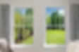 Two windows reveal a lush garden and green lawn with trees under a clear blue sky. Inside, patterned curtains and a beige chair are visible.