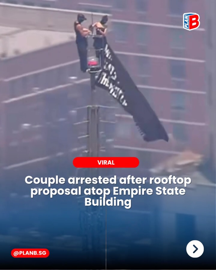 Couple on Empire State Building rooftop clings to a pole with a black flag, amid news text about a proposal and arrest.
