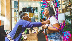 A Limin' Professionals coordinator helping a guest fit her teal and pink feathered Trinidad Carnival costume backpack at the numbered distribution station