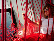 A woman in sunglasses and a white tee relaxes in a red rope hammock on a Virgin Voyages ship balcony, smiling out at the ocean in golden evening light.
