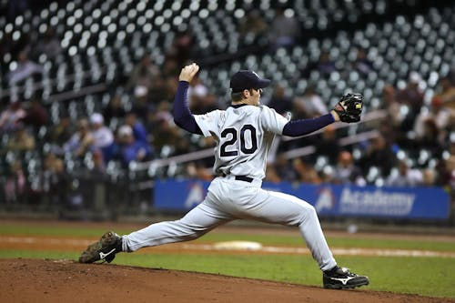 A baseball pitcher in a gray uniform with the number 20 is captured mid-pitch on the mound. His right arm is extended back, and his left leg is fully stretched forward as he throws the ball. The background features a stadium with spectators and empty seats.
