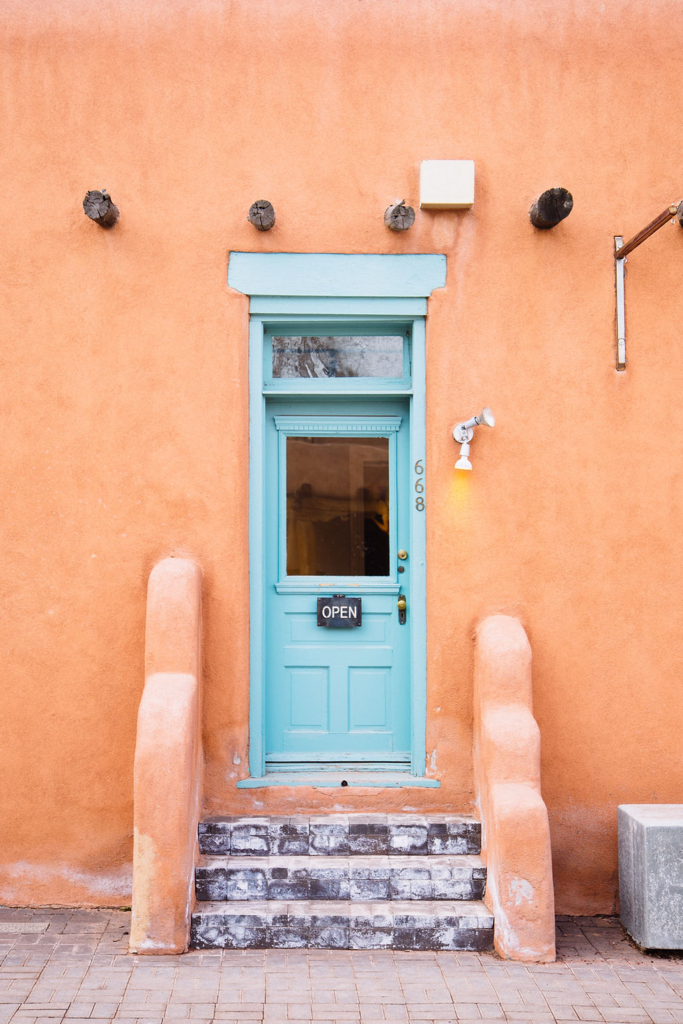 Teal door with "OPEN" sign, surrounded by orange adobe walls, steps leading to the entrance.