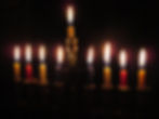 Background is black as night. Foreground is weakly lit by candles to show a golden outline of a kinara for Kwanza. All 9 candles are lit. The leftmost candel is yellow followed by red, then yellow, then dark purple, then the higher, center most candle which is yellow, then the rest of the candles starting with purple, to yellow, red, and yellow.