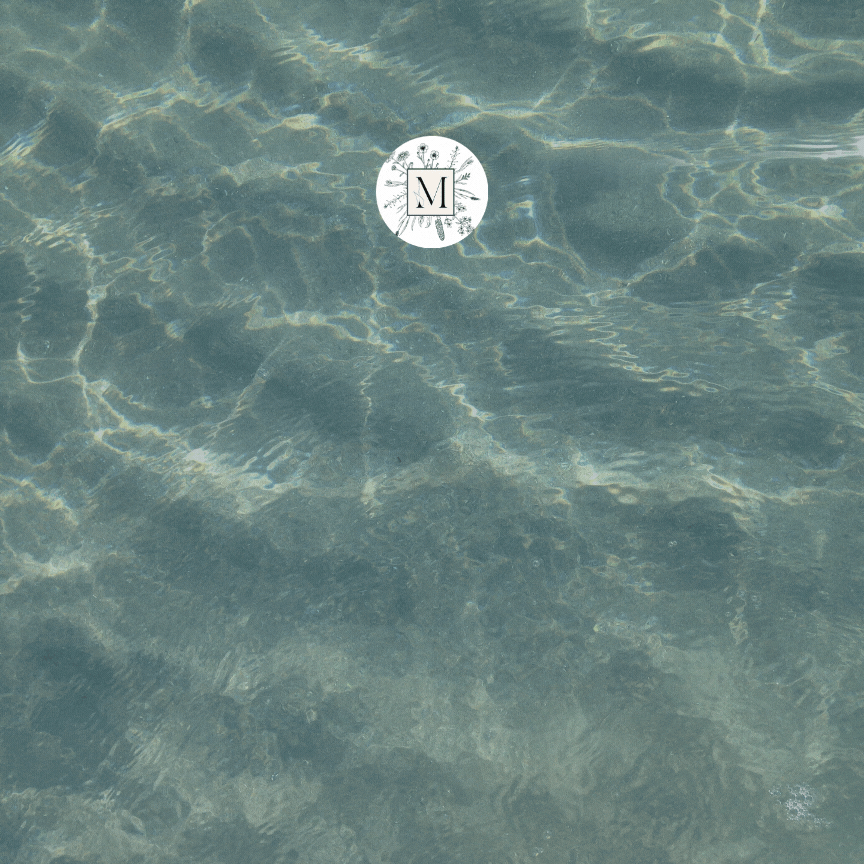 Clear blue water with sunlit ripples. A white circular logo featuring a central "M" and floral design floats on the surface, conveying serenity.