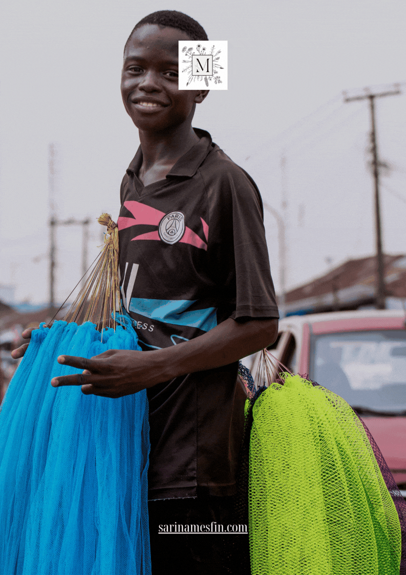 Smiling person holds blue and green net bags, wearing a black T-shirt with a logo. Background shows a car and overcast sky.
