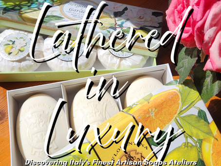 Soap boxes with citrus designs and white bars sit beside pink roses. Text reads "Lathered in Luxury" and "Discovering Italy's Finest Artisan Soaps Ateliers."