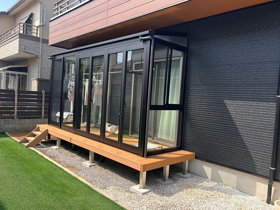 モダンな住宅の外観。黒いガラス張りのサンルームと木製デッキがあり、室内に洗濯物が干され、手入れされた芝生が広がる。