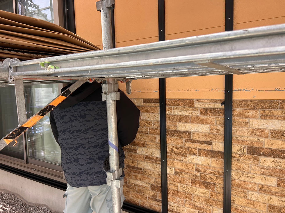 足場にいる作業員が建物の壁を施工中。壁はベージュと茶色のタイル模様で、オレンジ色の背景が見える。作業用ジャケット着用。