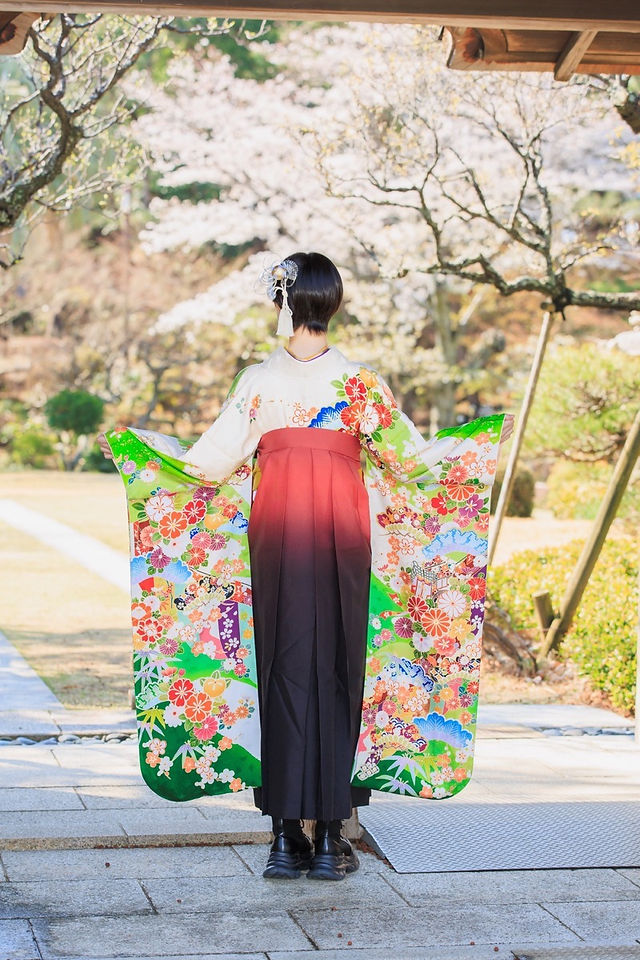 満開の桜の下、旧吉田茂邸で美しく着飾った姉妹が笑顔で並ぶ卒業記念の家族写真。春のやわらかな光に包まれた、家族の温かな思い出を写した一枚。