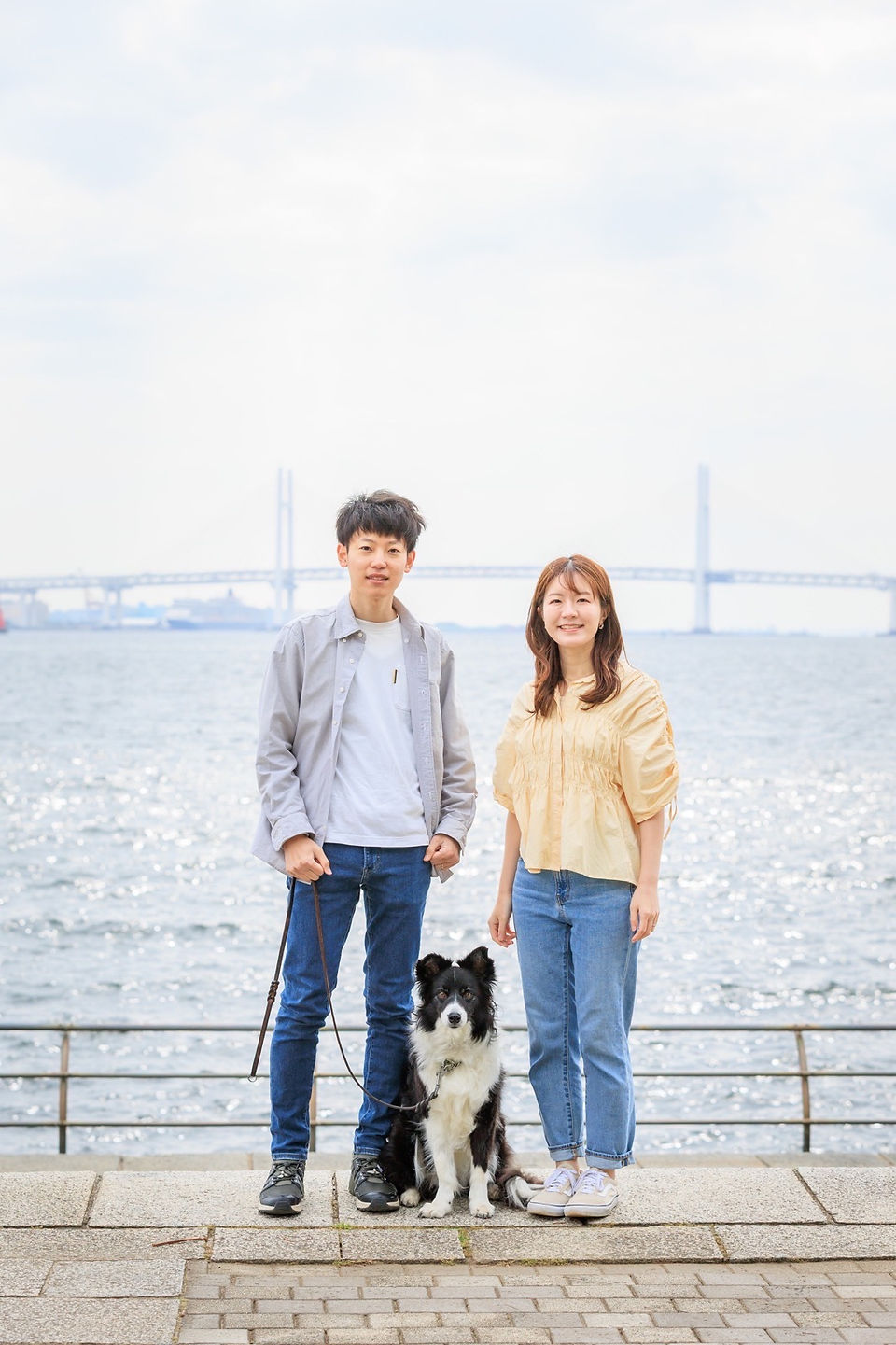 横浜・臨港パークでのペット撮影。シニア犬と飼い主夫婦が笑顔で寄り添う、自然光に包まれた家族写真。出張撮影ならではのリラックスした雰囲気が伝わる一枚