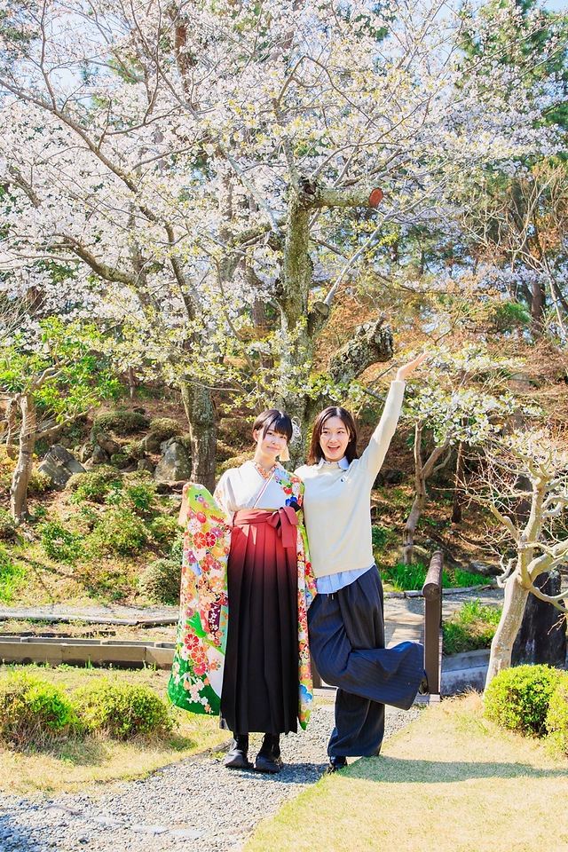 満開の桜の下、旧吉田茂邸で美しく着飾った姉妹が笑顔で並ぶ卒業記念の家族写真。春のやわらかな光に包まれた、家族の温かな思い出を写した一枚。