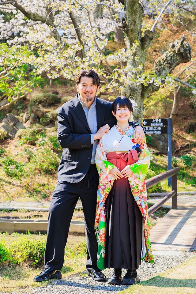 満開の桜の下、旧吉田茂邸で美しく着飾った姉妹が笑顔で並ぶ卒業記念の家族写真。春のやわらかな光に包まれた、家族の温かな思い出を写した一枚。
