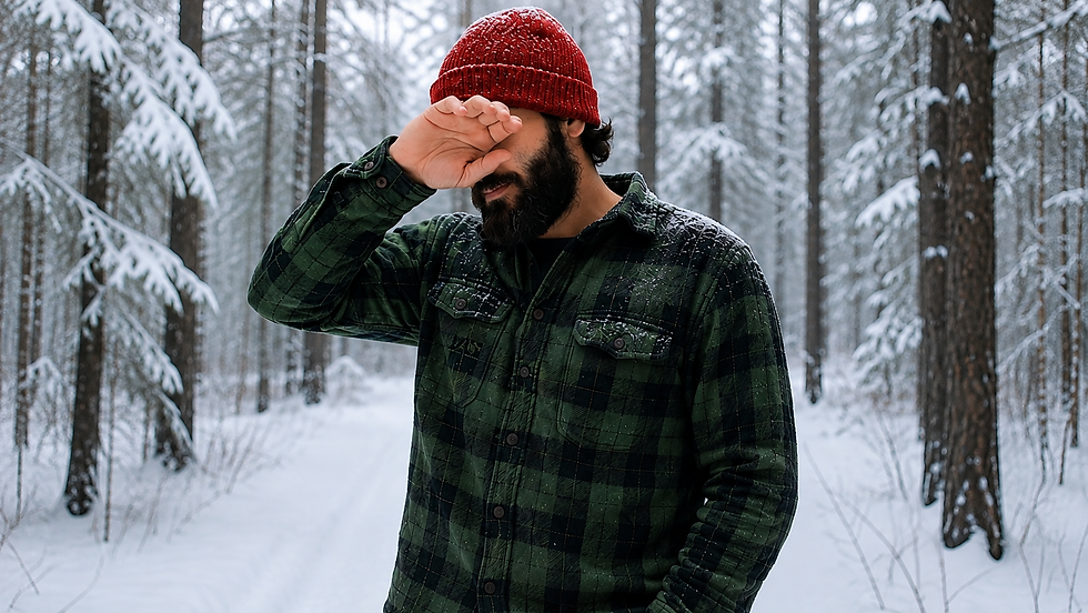 A bearded man wearing a red beanie and green plaid shirt wipes sweat from his face while standing in a snowy forest