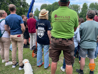 Gruppe von Menschen steht auf einer Wiese bei einer Outdoor-Veranstaltung, dem Entenrennen 2024 des Onkologischen Forums Celle e.V.. Im Mittelpunkt eine Frau mit Strohhut, SicherheitsCelle-T-Shirt und kleinem weißen Hund namens Jeanny an der Leine. Daneben ein Mann von hinten mit grünem Mylius-Garten-Landschafts-BauShirt.