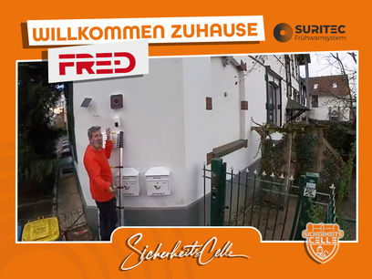 Björn Emanuelsson von SicherheitsCelle montiert ein FR.ED Frühwarnsystem an einer Hauswand neben zwei Briefkästen. Text: Willkommen Zuhause FR.ED, Logos von Suritec und SicherheitsCelle.