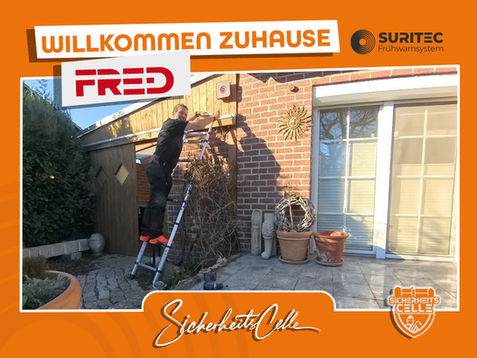 Björn Emanuelsson von SicherheitsCelle montiert ein FR.ED Frühwarnsystem an einer Hausfassade aus rotem Klinker. Text: Willkommen Zuhause FR.ED, Logos von Suritec und SicherheitsCelle.