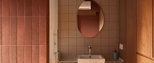 Moody washroom vanity nook with a round mirror, glowing globe sconce, terracotta wall tiles, and rich wood cabinetry.