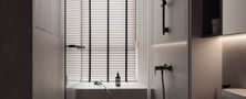 Sleek modern bathroom featuring dark floating vanity, wall-mounted toilet, long layout, and deep soaking bathtub set against window blinds.